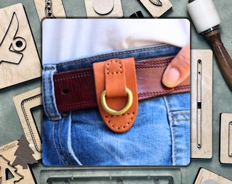 Könnte beinhalten: Ein brauner Ledergürtel mit einer kleinen, rechteckigen Ledertasche. Die Tasche hat einen Messingring und ist mit Nähten am Gürtel befestigt. Der Gürtel wird über einer blauen Jeans getragen.