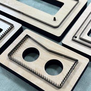 May include: Close-up of several wooden die-cut templates with black edges. The templates have various shapes, including rectangles and a design with two circular cutouts and a jagged edge. The wood has a light, natural color.