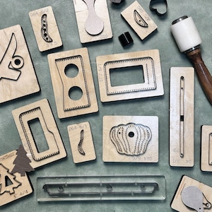 May include: A collection of various wooden leathercraft cutting dies, including shapes like rectangles, ovals, and organic forms. Some dies have numbers and letters printed on them, such as "064 6/2" and "345". A mallet is also present.