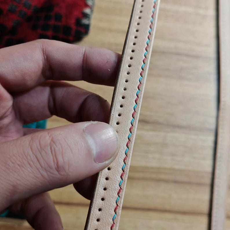 May include: Close-up of a light brown leather strap with a row of evenly spaced holes and a decorative stitch in red, blue, and turquoise.