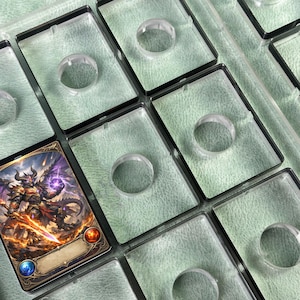 Peut inclure: Gros plan sur des protège-cartes en plastique transparent avec un cadre noir. Une carte est visible, avec une illustration fantastique d'une créature ressemblant à un dragon. Les supports ont une indentation circulaire au centre.