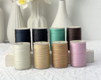 May include: Eight cylindrical spools of thread arranged in two rows. The top row features dark blue, black, mint green, and dark brown spools. The bottom row includes off-white, beige, tan, and light pink spools, all wound with smooth cord.