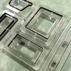May include: Clear acrylic molds with black rubber seals. The molds are rectangular and square, with various shapes and sizes. The image shows a collection of molds, likely for crafting or industrial use, arranged on a textured surface.