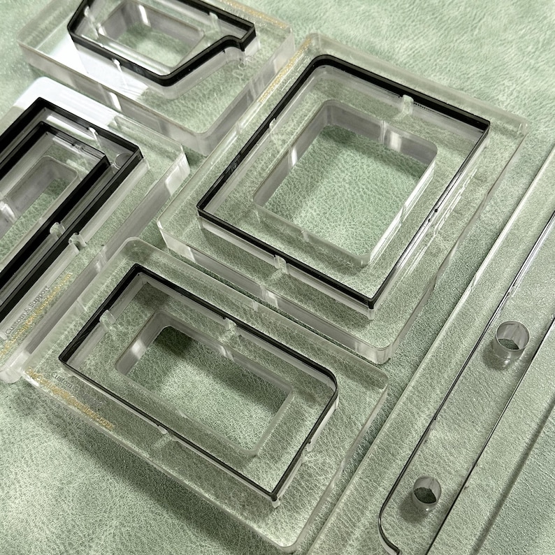 May include: Clear acrylic molds with black rubber seals. The molds are rectangular and square, with various shapes and sizes. The image shows a collection of molds, likely for crafting or industrial use, arranged on a textured surface.