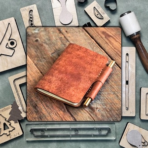 May include: A brown leather notebook with a pen holder and a gold pen, on a wooden surface. Various leather crafting tools surround the notebook, including wooden stamps and a mallet.