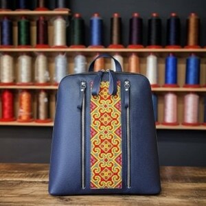 May include: A navy blue backpack with a central panel featuring a colorful, ornate pattern in red, yellow, and pink. The backpack has two vertical zippered pockets and a top handle. It sits on a wooden surface.