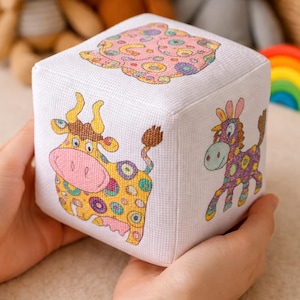Può includere: Un cubo di stoffa bianco con illustrazioni di cartoni animati colorati di una mucca, un cavallo e un orso. Il cubo è tenuto tra le mani di qualcuno, con altri peluche visibili sullo sfondo.