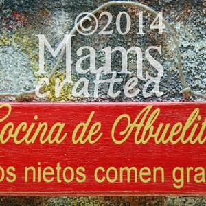 May include: Red wooden sign with white text that reads "Cocina de Abuelita Los nietos comen gratis". The sign has a rope hanger and is hanging on a textured gray background.
