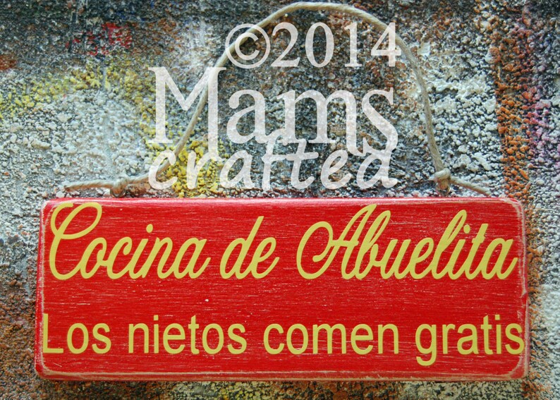 May include: Red wooden sign with white text that reads "Cocina de Abuelita Los nietos comen gratis". The sign has a rope hanger and is hanging on a textured gray background.
