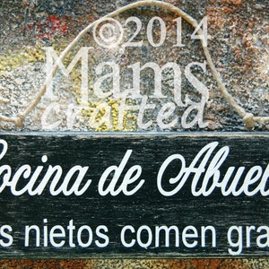 May include: A black wooden sign with white text that reads "Cocina de Abuela Los nietos comen gratis". The sign is hanging from a string and has a distressed look.