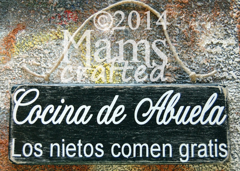 May include: A black wooden sign with white text that reads "Cocina de Abuela Los nietos comen gratis". The sign is hanging from a string and has a distressed look.