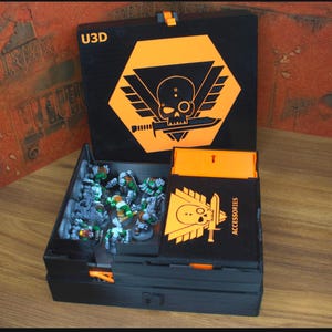 May include: Black storage box with orange accents, featuring a skull and sword emblem. The box is open, revealing miniature figures inside. The lid displays the text "U3D". A smaller box labeled "ACCESSORIES" is also visible.