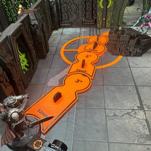May include: An orange game board accessory with keyhole-shaped cutouts, marked with measurements of 2", 3", and 32mm. The accessory is placed on a gray tiled surface, with miniature figures and a black structure in the background. The scene suggests a tabletop game.