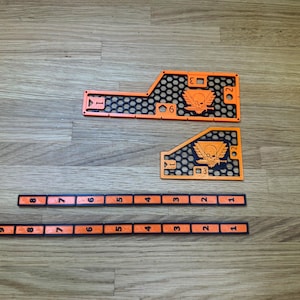 Op de afbeelding: Oranje en zwarte spelcomponenten, waaronder genummerde markeringen en gevormde stukken met een schedel- en vleugel-embleem. De stukken zijn gerangschikt op een houten oppervlak, met een honingraatpatroon.