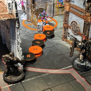 May include: A tabletop gaming scene featuring miniature figures and terrain. The scene includes several figures, orange and black tokens marked with "25", and detailed building structures. The figures are in various poses, suggesting an active game.