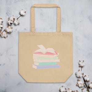 Könnte beinhalten: Eine beige Canvas-Tragetasche mit einer bunten Illustration eines Stapels Bücher. Das oberste Buch ist geöffnet und zeigt ein rosafarbenes Innere.