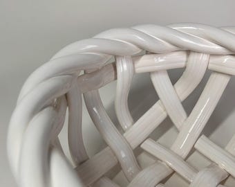 May include: Close-up of a white ceramic basket with a woven design. The basket's rim features a twisted rope-like detail, and the body has a lattice pattern. The glossy finish reflects light, highlighting the basket's texture and form.