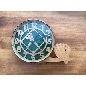 Peut inclure: Une horloge ronde avec un cadran vert et une bordure dorée. L'horloge présente une silhouette de joueur de baseball et un gant de baseball en bois attaché sur le côté. L'horloge est posée sur une surface en bois.