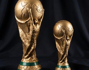 May include: Two gold-colored FIFA World Cup trophy replicas of different sizes. Each trophy features a stylized human figure holding up a globe, set upon a circular green base with the embossed text FIFA WORLD CUP visible on the front.