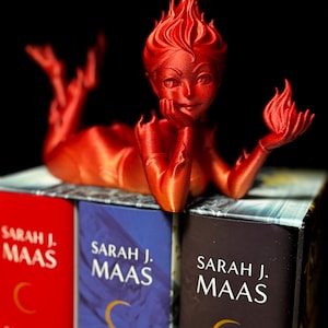May include: A red 3D-printed figurine of a woman with flame-like hair, reclining on a stack of books. The books have the author's name, Sarah J. Maas, and the titles Crescent City and other titles. The figurine is holding a flame in one hand.