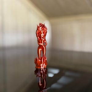 May include: A small, red-orange figurine of a person engulfed in flames. The figure is standing on a base of flames, with the head and upper body also shaped like flames. The figurine is highly reflective, with a smooth, glossy finish.
