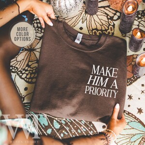 May include: A brown sweatshirt with the text "MAKE HIM A PRIORITY" in white. The sweatshirt is folded and held by hands. The words "MORE COLOR OPTIONS" are in a circle in the upper left corner.