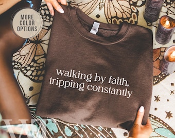 May include: A brown sweatshirt with the text "walking by faith, tripping constantly" in white. The sweatshirt is held by hands, with a decorative cloth and candles in the background. The words "More Color Options" are in a circle.