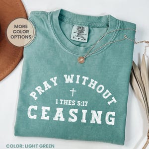 May include: Light green t-shirt with the text "PRAY WITHOUT CEASING" and a cross graphic. The shirt also features the text "1 THES 5:17". A gold necklace with a pendant is draped on the shirt. The shirt is labeled "Comfort Colors".