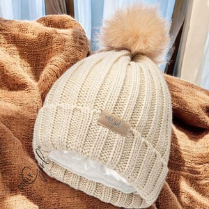 May include: Cream-colored knit beanie with a faux fur pom-pom. The hat features a ribbed design and a small rectangular tag with the text "Curly Girl". The interior lining appears to be a satin material. The hat is resting on a brown blanket.
