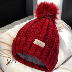May include: A vibrant red knit hat with a large, fluffy pom-pom on top. The hat features a ribbed knit design and a small tag that reads "Curly Girl". The hat is resting on a white pillow.