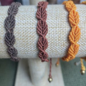 Pu&ograve; includere: Tre braccialetti intrecciati in marrone, arancione e rosso, esposti su un supporto rivestito in tessuto. I braccialetti sono realizzati con un motivo a forma di foglia.