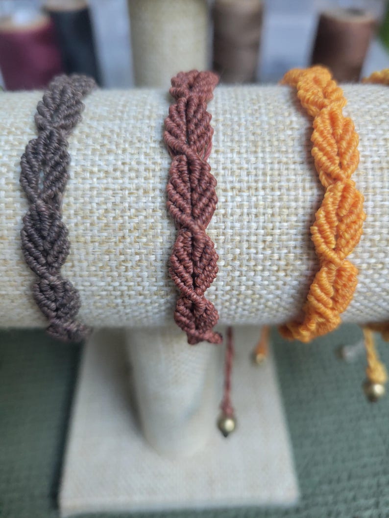 Pu&ograve; includere: Tre braccialetti intrecciati in marrone, arancione e rosso, esposti su un supporto rivestito in tessuto. I braccialetti sono realizzati con un motivo a forma di foglia.