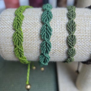 Pu&ograve; includere: Tre braccialetti dell'amicizia verdi con motivi a foglia, esposti su un supporto in tessuto beige. Ogni braccialetto ha un perno dorato alla fine.