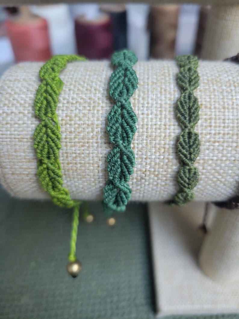 Pu&ograve; includere: Tre braccialetti dell'amicizia verdi con motivi a foglia, esposti su un supporto in tessuto beige. Ogni braccialetto ha un perno dorato alla fine.