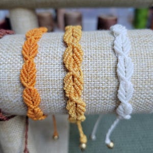 Pu&ograve; includere: Tre braccialetti dell'amicizia in arancione, giallo e bianco. I braccialetti sono realizzati con un cordoncino annodato e hanno una piccola perla all'estremit&agrave;.