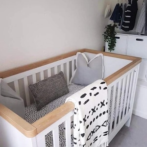 May include: A white cot with a wooden frame and a black and white patterned sheet. A white blanket with black geometric designs is draped over the side of the cot.
