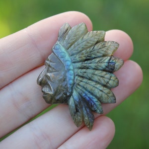 May include: A carved labradorite gemstone pendant featuring a Native American chief wearing a headdress. The gemstone has a dark gray base with iridescent blue and green flashes.