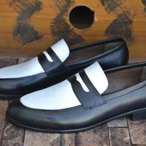 May include: A pair of black and white leather loafers. The shoes have a classic design with a black body and a white vamp and strap. The shoes are displayed on a brown, textured surface.