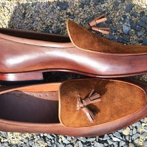Peut inclure: Une paire de mocassins en cuir marron avec des accents en daim et des glands. Les mocassins ont une semelle en cuir marron et un talon en cuir marron.