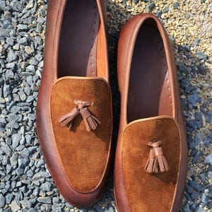 Peut inclure: Une paire de mocassins en cuir marron avec des glands en daim. Les mocassins ont une semelle en caoutchouc et sont pos&eacute;s sur un lit de gravier.