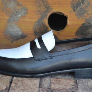May include: A black and white leather loafer. The shoe features a black body with a white vamp and a black strap across the top. The sole is black, and the shoe has a low heel. The background is a wooden surface.