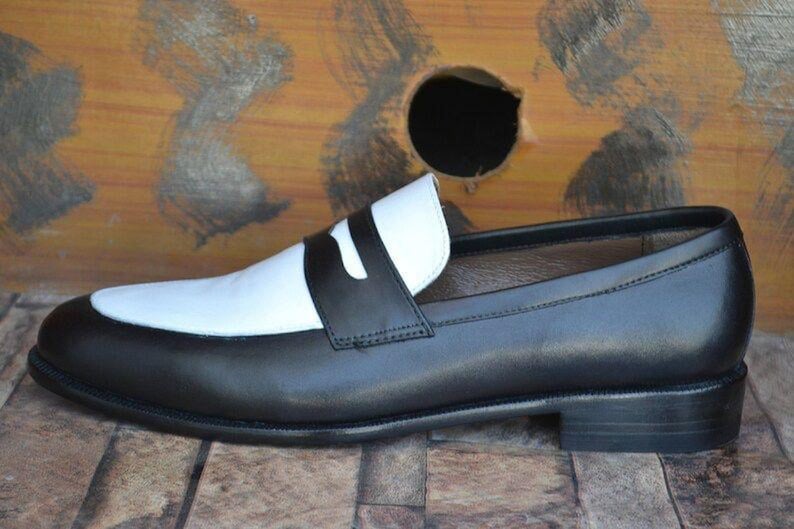 May include: A black and white leather loafer. The shoe features a black body with a white vamp and a black strap across the top. The sole is black, and the shoe has a low heel. The background is a wooden surface.