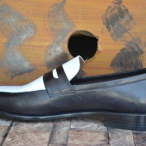 May include: A black and white leather loafer shoe. The shoe features a black body with a white vamp and strap. The shoe is set against a wooden background.
