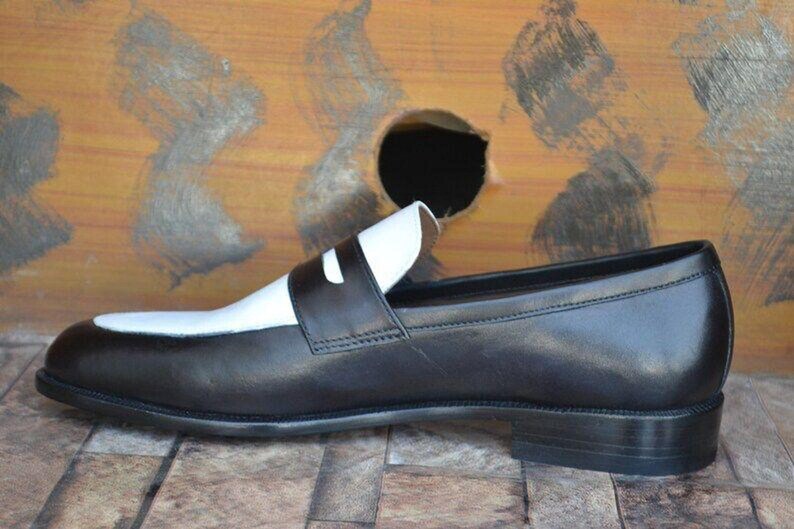 May include: A black and white leather loafer shoe. The shoe features a black body with a white vamp and strap. The shoe is set against a wooden background.