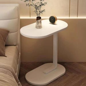 May include: A white bedside table with an oval top and base, connected by a curved leg. A small gold-rimmed clock and a vase with greenery sit on the table. The table is next to a bed with a white headboard.