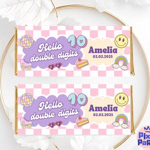 Puede incluir: Dos envoltorios de barras de caramelo con un patrón de cuadros rosas y blancos. Cada envoltorio tiene el texto "Hello double digits" y "Amelia 02.02.2025". Los envoltorios también presentan un patín, una bola de discoteca, un arcoíris y un pastel.