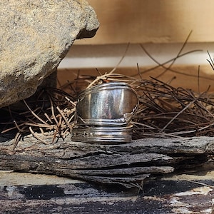 May include: A handcrafted silver ring made from repurposed silverware. The ring features a spoon bowl design on top and a decorative band. The ring is displayed on a piece of weathered wood, with a natural background.