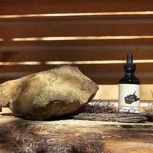 Puede incluir: Una botella de vidrio verde oscuro con un cuentagotas, una etiqueta blanca con un logotipo negro y el texto "Aura". Una roca grande y texturizada se encuentra junto a la botella. Los artículos se exhiben en una caja de madera.