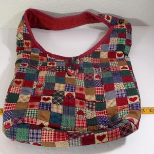 May include: A patchwork quilted shoulder bag with a red heart button closure. The bag is made of various fabrics in shades of red, green, blue, and brown.