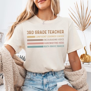May include: Cream-colored t-shirt with the text "3RD GRADE TEACHER" in brown, with colorful stripes below listing teacher qualities. The shirt is worn by a person in blue jeans.
